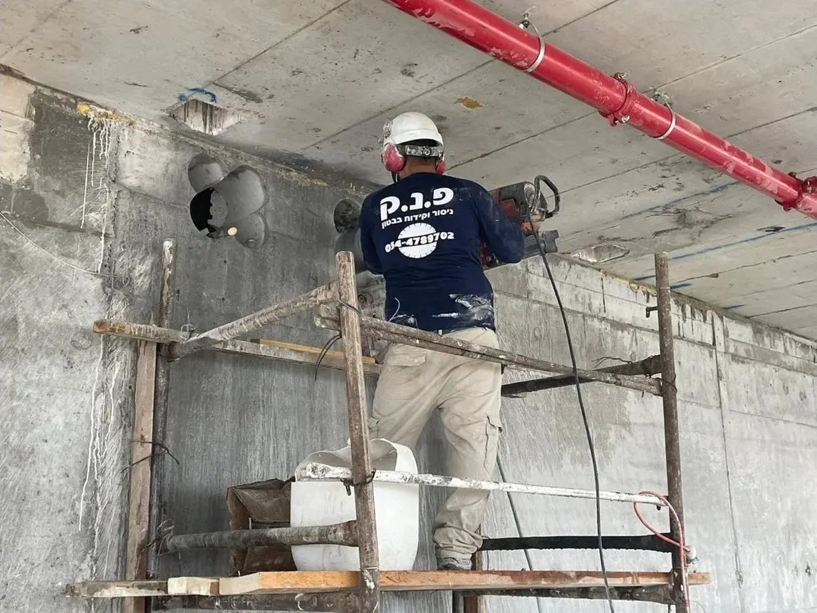 עובד פ.נ.ק מבצע קידוח בטון ביהלום באמצעות מקדחת ליבה על גבי פיגום
